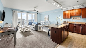 A modern living room with a sofa, coffee table, and dining table. There is a kitchen area with wooden cabinets and a large window. - SMOOTH SAILING-BEAUTIFULLY DECORATED, GULF VIEWS (Panama City Beach)
