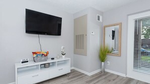 A modern living room with a flat-screen TV mounted on the wall, a white entertainment unit with a basket of snacks, a potted plant, and a large mirror. - Shopping Paradise | Free coffee + Parking (Kettering)