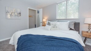 A bedroom with a bed, a bedside table with a lamp, a window with blinds, a door leading to a bathroom, and a wall-mounted artwork. - Shopping Paradise | Free coffee + Parking (Kettering)
