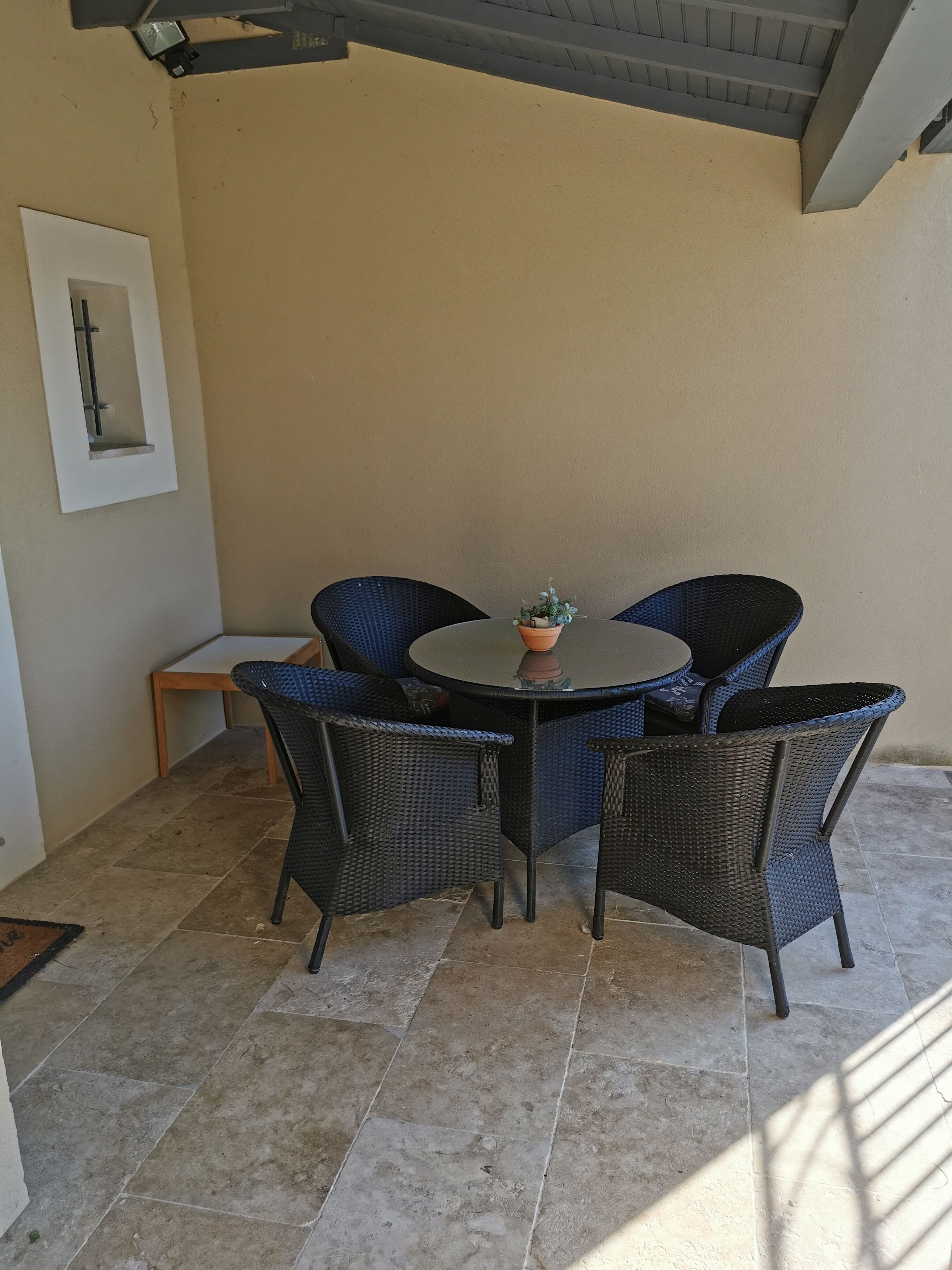 Une terrasse extérieure avec une table ronde et quatre chaises en osier, une petite plante en pot et un tableau encadré accroché au mur.