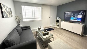 Una sala de estar moderna con un sofá gris, una mesa de centro de vidrio, un televisor sobre un mueble bajo y una ventana con persianas.