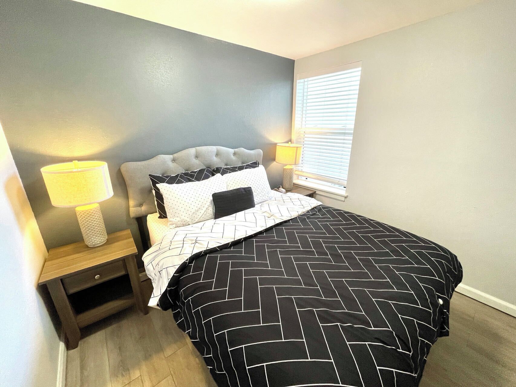 A bedroom with a tufted headboard, a bed with a black and white patterned bedspread, two bedside lamps, and a window with blinds.