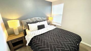 A bedroom with a tufted headboard, a bed with a black and white patterned bedspread, two bedside lamps, and a window with blinds.