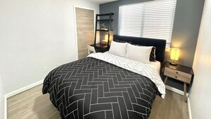 Un dormitorio con cama, mesitas de noche, un armario auxiliar, una ventana con persianas y un estante fijado a la pared.