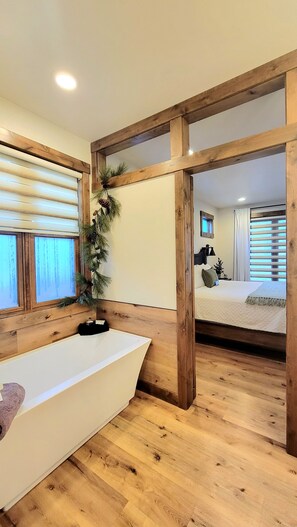 A room with a wooden floor, a white bathtub, and a bed with a white headboard.