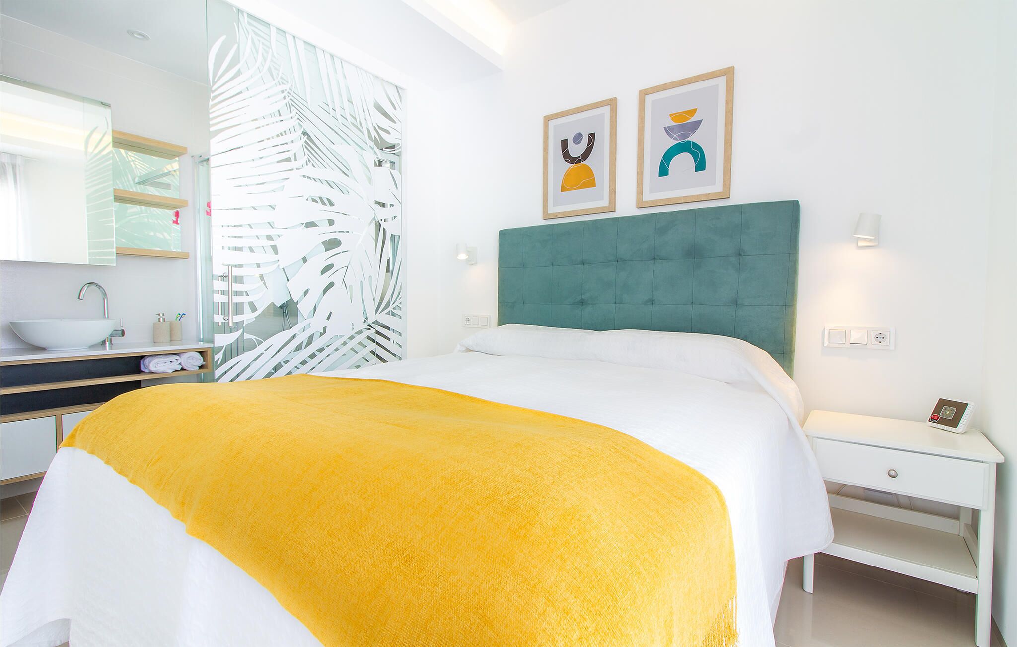 Une chambre d’hôtel moderne dotée d’un grand lit, d’une table de chevet, d’une commode, d’une salle de bain avec un lavabo et un miroir, et d’œuvres d’art abstraites accrochées au mur.