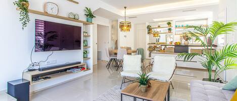 A modern living room with a flat-screen TV, wooden shelves, and a dining area with a chandelier.