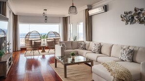 A modern living room with a large sofa, a glass coffee table, wicker chairs, and a view of the ocean through large windows.