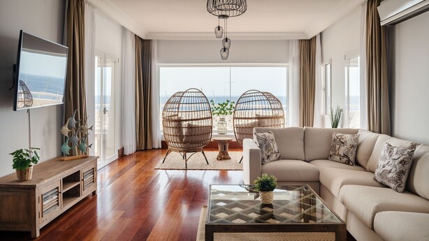 A modern living room with a large sofa, a glass coffee table, wicker chairs, and a TV on the wall.