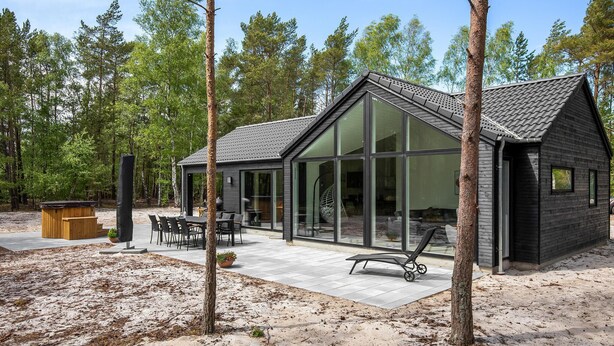 Una cabaña moderna con grandes ventanales, un patio con mobiliario exterior y un telón de fondo boscoso.