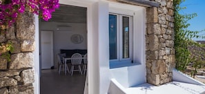 L’extérieur est en pierre, avec une porte blanche et une petite fenêtre. À l’intérieur, on trouve un coin repas avec une table et des chaises.
