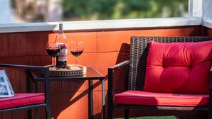 Una silla de mimbre con un cojín rojo, una mesita con una botella de vino y dos copas, y un cuadro en la mesita.