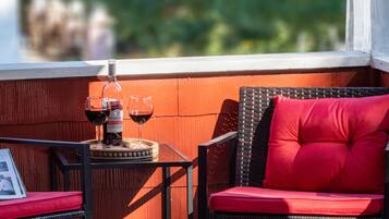 Une chaise en osier avec un coussin rouge, une petite table sur laquelle se trouvent une bouteille de vin et deux verres, et un tableau encadré posé sur la table.