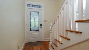 Un pasillo con piso de madera, una puerta blanca con panel de vidrio y barandal blanco con pasamanos de madera.