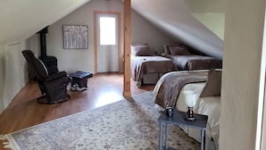 Un dormitorio acogedor en el ático, con techo inclinado, ventilador de techo, chimenea, dos camas con ropa de cama a juego, un sillón mecedor y una mesita de noche con lámpara.