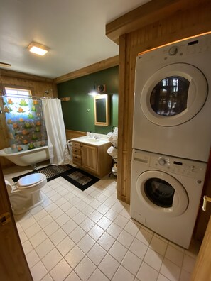 Uma casa de banho compacta com sanita, lavatório, chuveiro e uma máquina de lavar roupa empilhada com secadora.