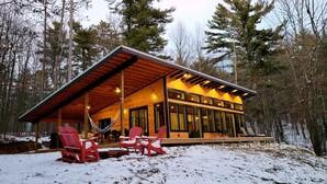 Una moderna baita con rivestimento in legno, ampie finestre e una zona salotto esterna coperta con sedie rosse.