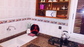 A kitchen with white tiles, a granite countertop, a sink, a stove, and a wooden shelf with dishes and kitchenware.