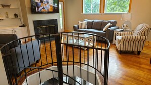 Un salon moderne doté d’une cheminée, d’un téléviseur fixé au mur, d’un escalier avec une rampe et offrant une vue sur l’extérieur grâce à de grandes fenêtres.