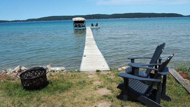 A dock extending into a calm lake with a boat at the end. There is a chair and a basket on the grassy shore. - The Bright and Breezy - Lakefront with Spa! (Frankfort)