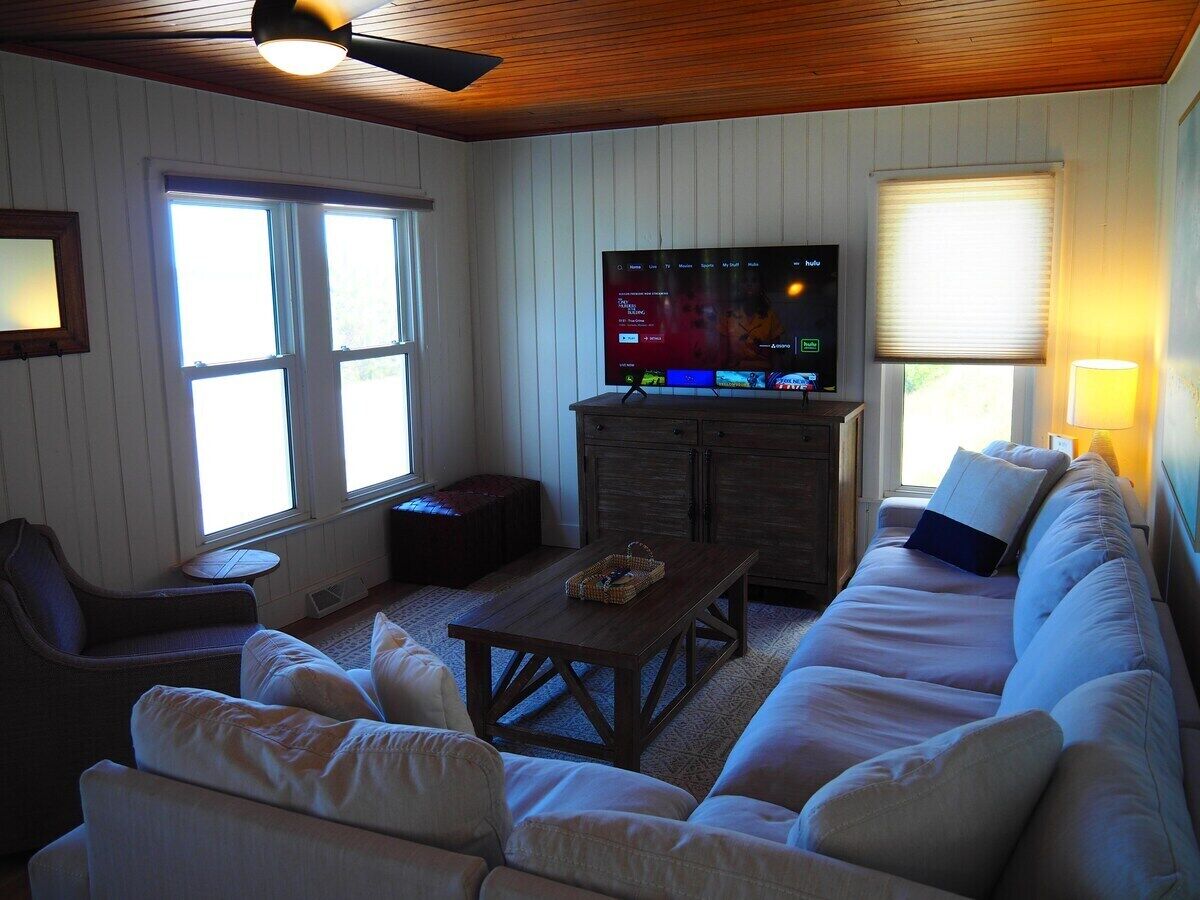 A cozy living room with a wooden ceiling, a large flat-screen TV, a sofa, a coffee table, and a window with blinds.