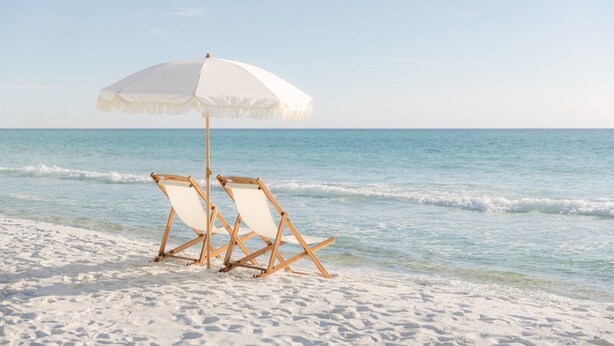 Two wooden beach chairs under a white umbrella on a sandy beach with clear blue water.