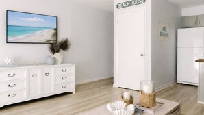 A room with a white dresser, a television displaying a beach scene, a door labeled "BEACH HOUSE", and a small basket with a candle.