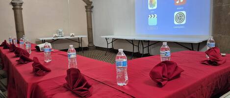 A conference room with tables covered in red tablecloths, water bottles, and red napkins. A projector screen displays icons for music, photo, and settings.