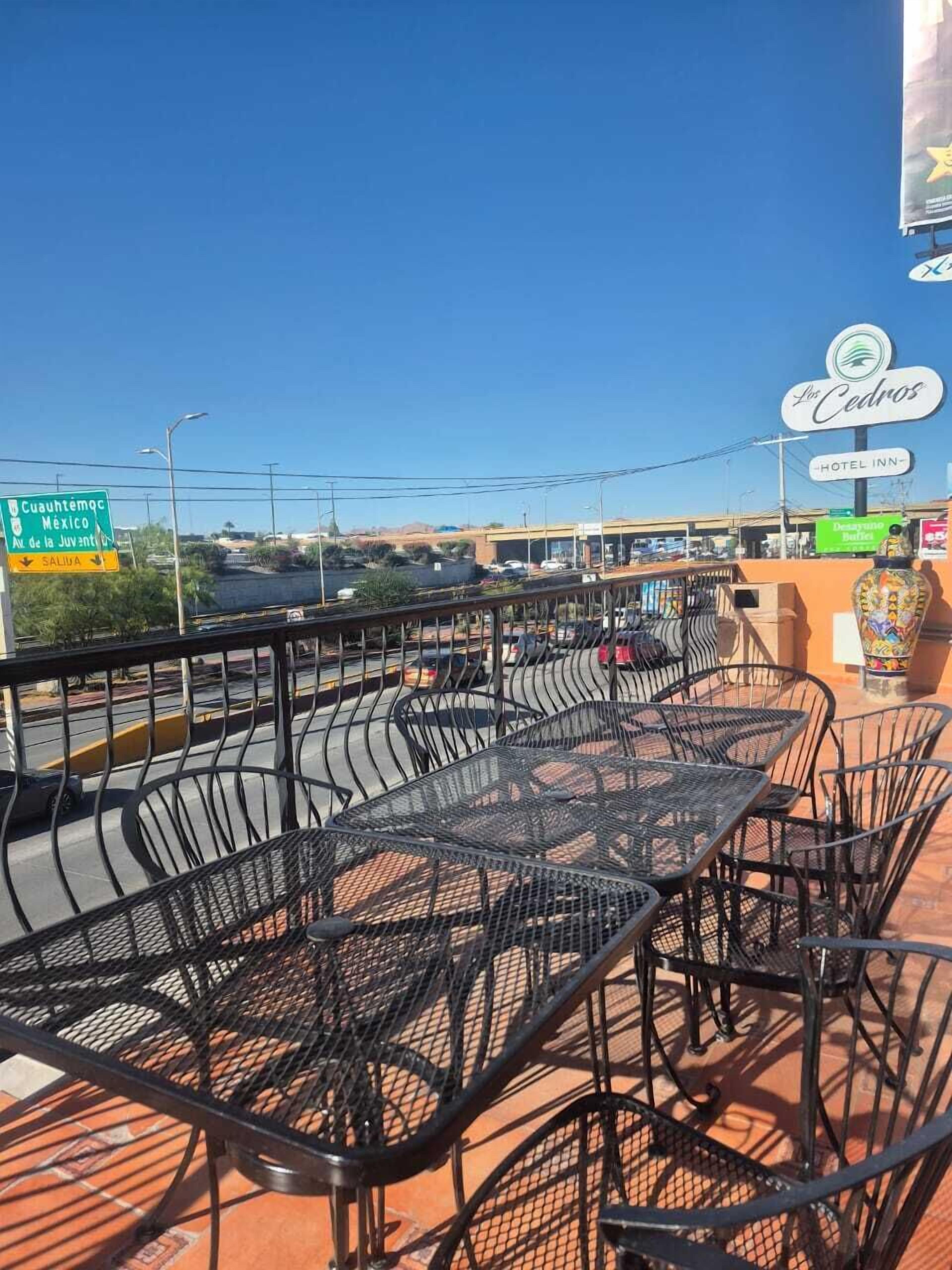 Outdoor seating with metal chairs and tables, a clear sky, and a sign for 'Los Cedros Hotel Inn' in the background.