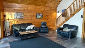 A cozy living room with wooden walls, a black leather sofa, a recliner, and a staircase.