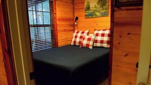 A wooden cabin room with a bed, checkered pillows, a painting, and a window with blinds. - Modern Private Cabin, Fishing, Shooting Range, Fire Pit, 4WD Trails & Canoeing (Bridgeport)