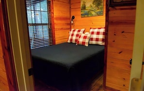 Una camera in una baita di legno con un letto, cuscini a quadretti, un quadro e una finestra con persiane.