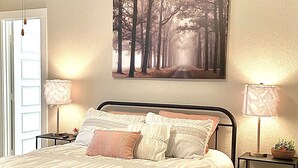 Una cama bien hecha con edredón blanco y varios almohadones, flanqueada por dos mesitas de noche con lámparas, una de ellas con una planta. Un cuadro de un sendero forestal cuelga sobre la cama.