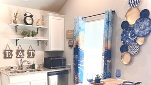 Cocina compacta con armarios blancos, un microondas y una pequeña isla con taburetes. Hay platos decorativos en la pared y una ventana con cortinas azules.