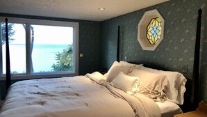 Une chambre à coucher avec un grand lit, une table de chevet, une commode en bois et une fenêtre donnant sur l’océan.