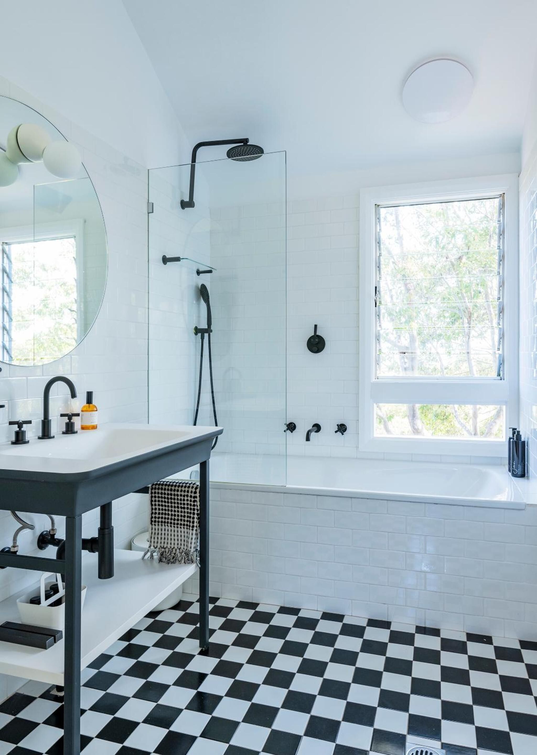 Un baño moderno con piso a cuadros, un espejo grande, un lavamanos con grifo, una ducha con mampara de vidrio y una ventana que proporciona luz natural.