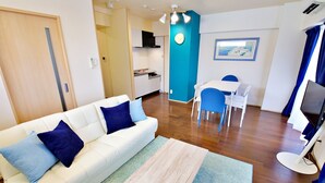 A modern living room with a white sofa, blue cushions, a wooden coffee table, and a dining area with a white table and blue chairs. - Rave K 201 (Naha)