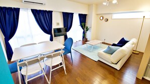 A living room with a white sofa, blue curtains, a flat-screen TV, a dining table with chairs, and a potted plant. - Rave K 201 (Naha)