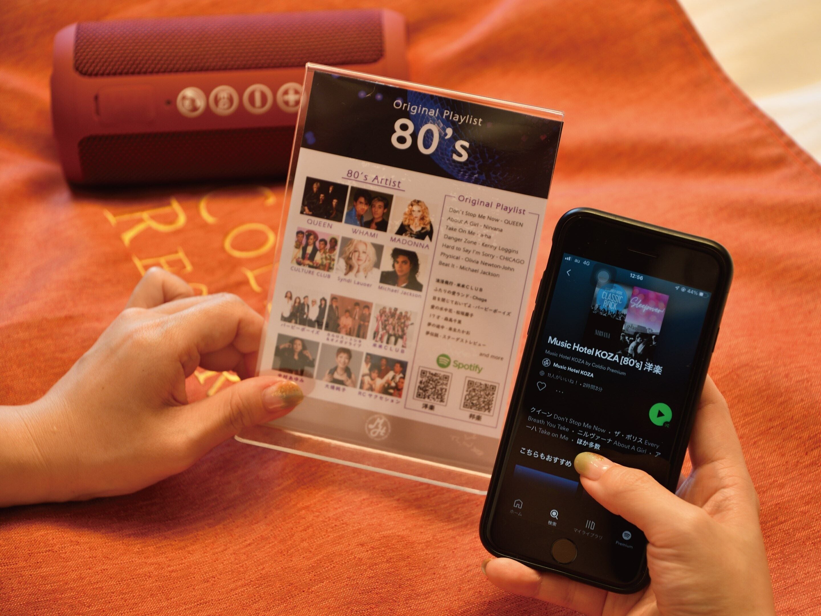 A person holding a smartphone displaying a music playlist titled "Music Hotel KOZA [80's]".