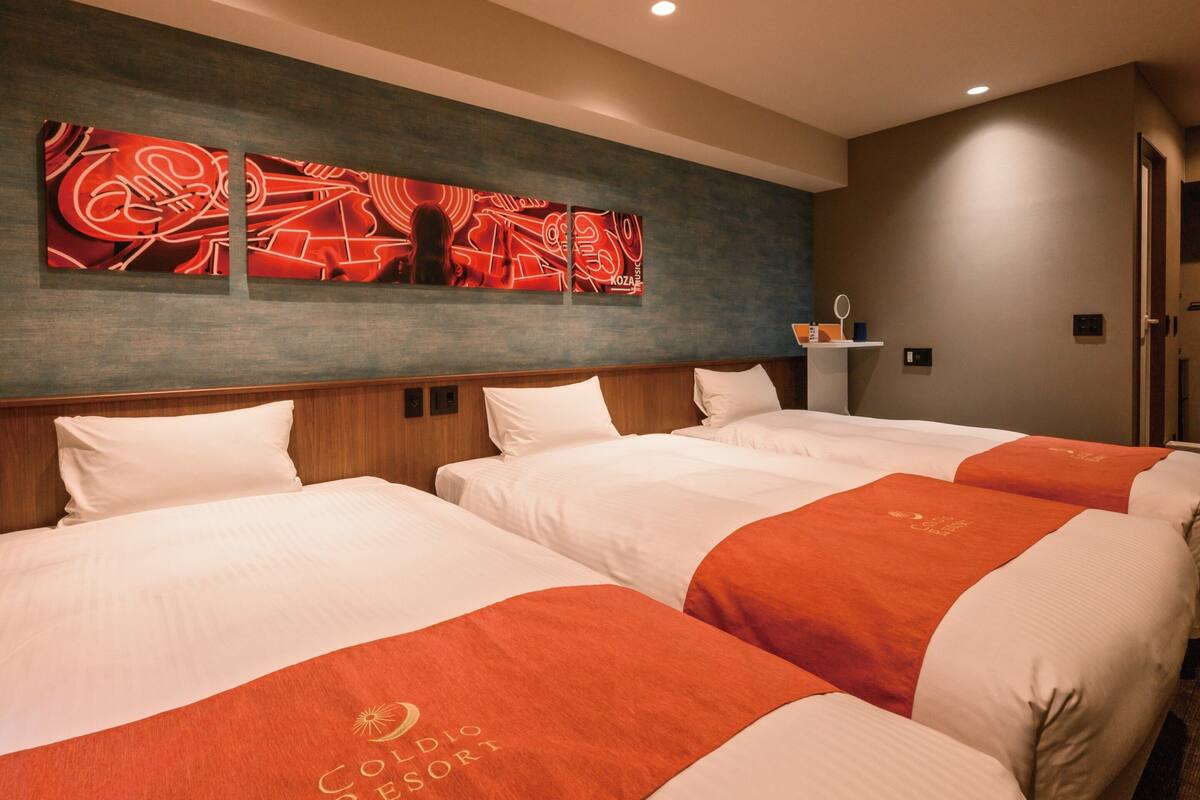 A hotel room with three beds, each with a red and white bedspread. There is a wall art piece above the beds and a small shelf with toiletries.