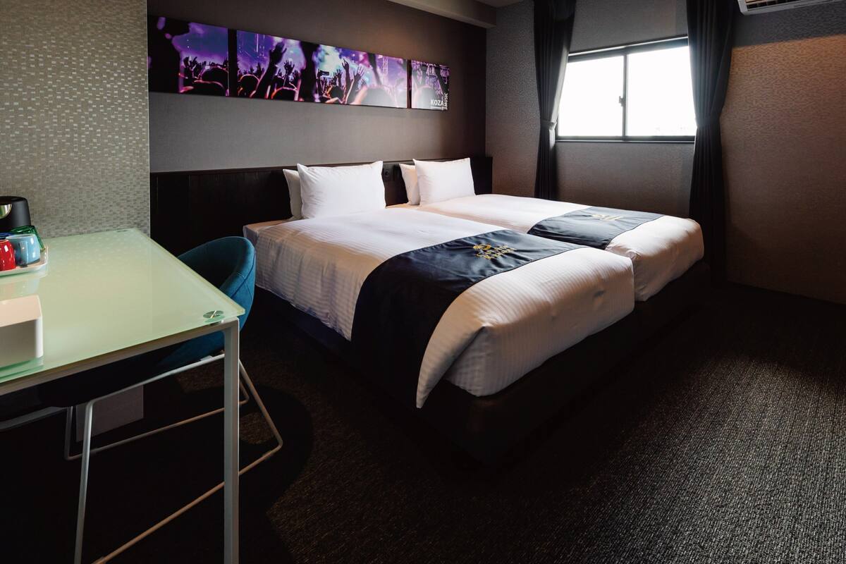 A hotel room with a bed, a desk, and a wall-mounted TV displaying a concert.