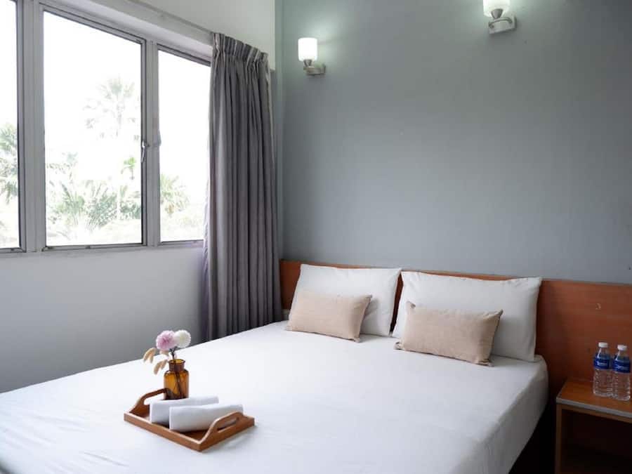 Una habitación de hotel con una cama, una mesita de noche con botellas de agua, una bandeja con toallas y un florero, y una ventana con cortinas.