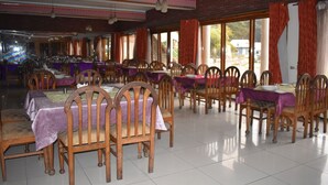 Uma sala de jantar espaçosa com várias mesas e cadeiras, janelas amplas com cortinas e piso de azulejos.