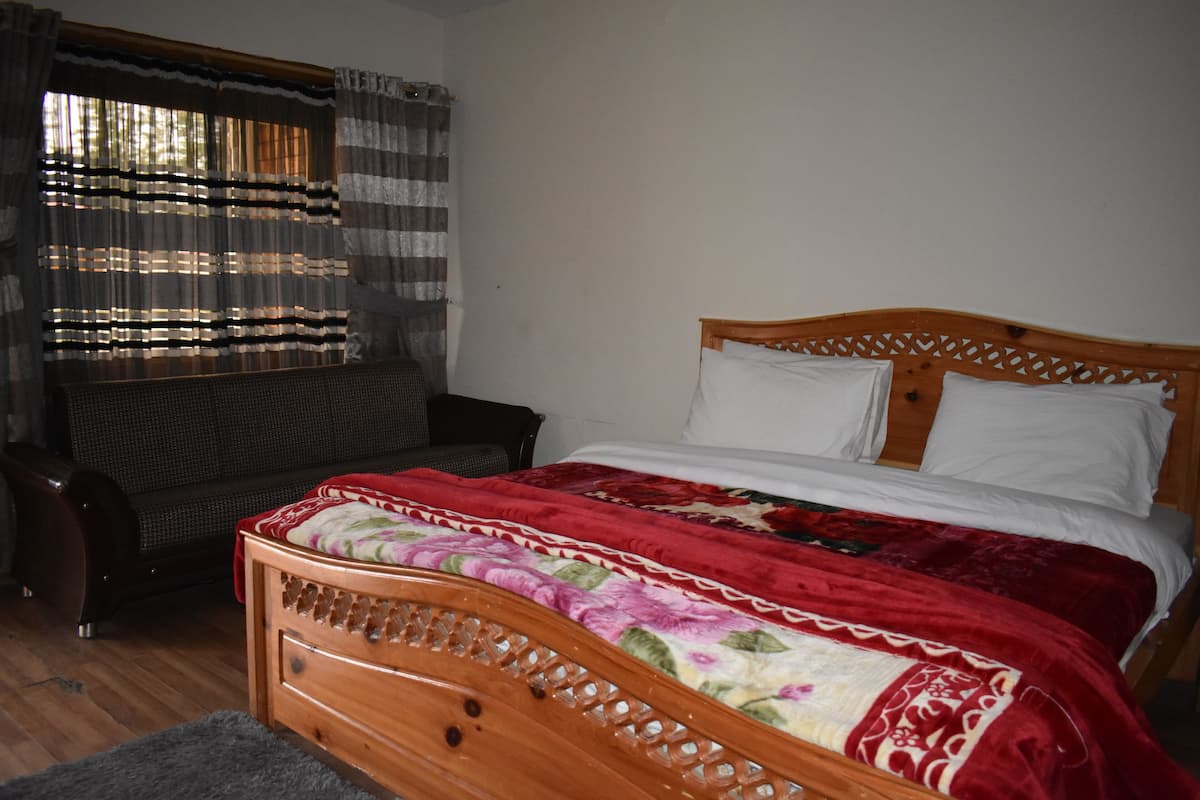 A bedroom with a wooden bed, a red and white floral bedspread, a grey rug, a wicker chair, and a window with striped curtains.
