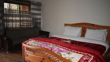 Un dormitorio con una cama de madera, una colcha floral roja y blanca, una alfombra gris, una silla de mimbre y una ventana con cortinas a rayas.
