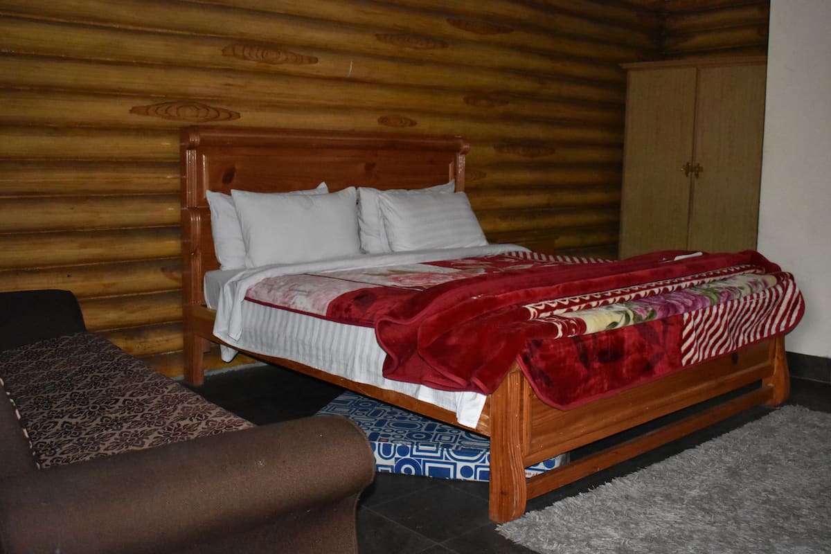 A wooden bed with white and red bedding, a patterned rug, and a wardrobe in a room with wooden walls.