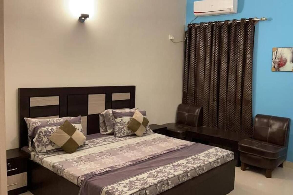 A bedroom with a bed, a desk, two chairs, a wall-mounted air conditioner, and a curtain.