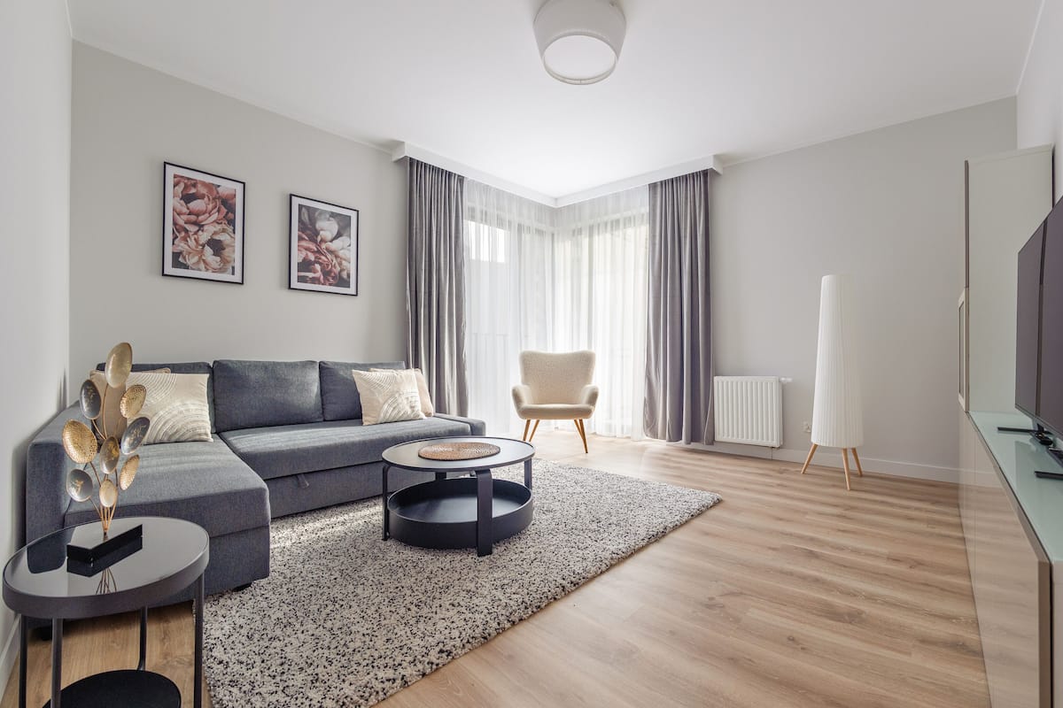 Una sala de estar moderna con un sofá gris, una mesa redonda de centro, una mesita auxiliar, una lámpara de pie alta, un televisor y un radiador.