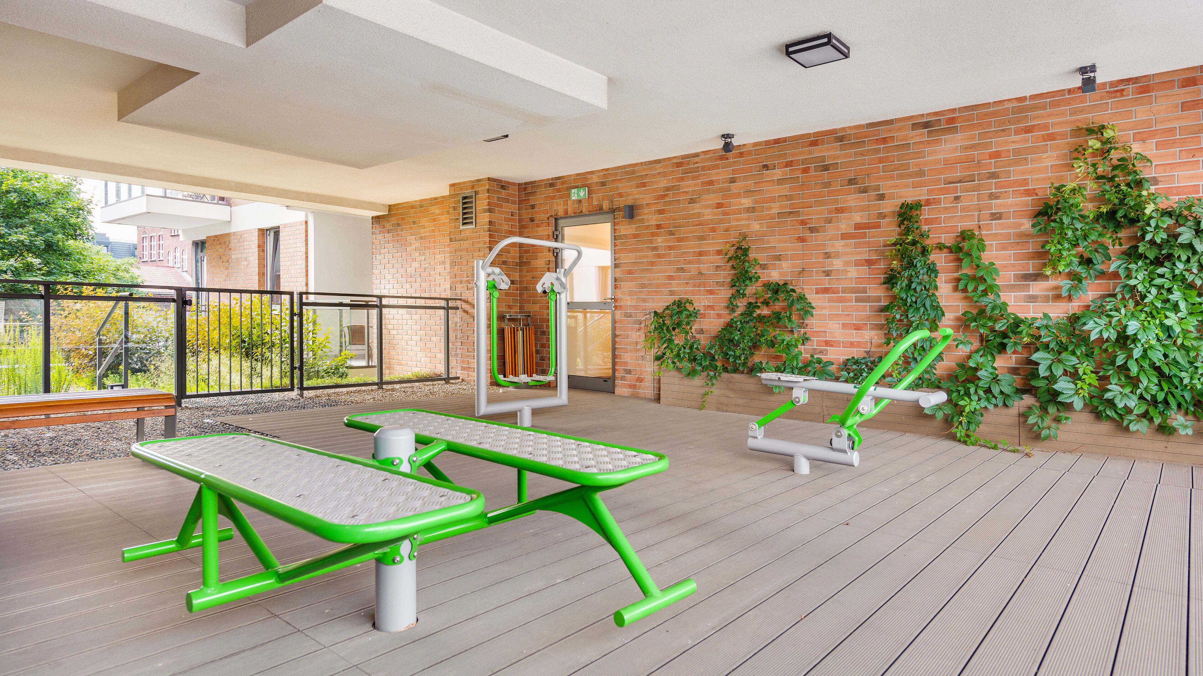 Un espacio exterior de acondicionamiento físico con equipamiento de ejercicio, una terraza de madera y un muro de ladrillo con vegetación.