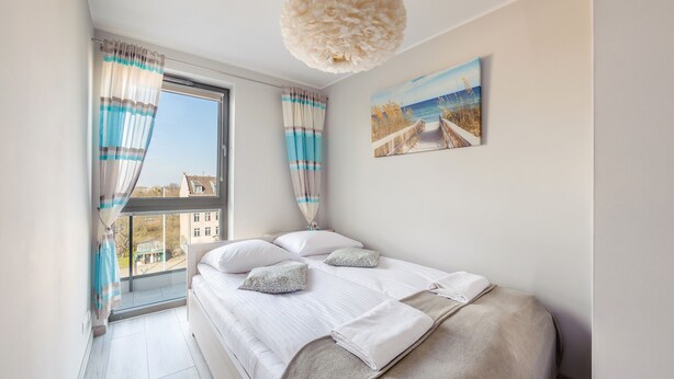 Een slaapkamer met een groot raam, een bed met witte lakens en een schilderij van een strand.